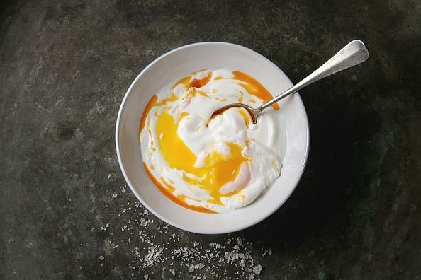 Poached egg on yogurt sauce with spicy olive oil, salt, spoon served in white plate over old dark metal background. Top view, copy space. Vegetarian healthy eating